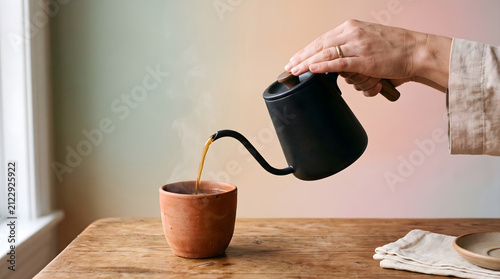 Hand pours hot coffee from a matte black gooseneck kettle into a rustic ceramic mug resting on a weathered wooden table inside a cozy room with soft ambient light creating a warm morning atmosphere.
