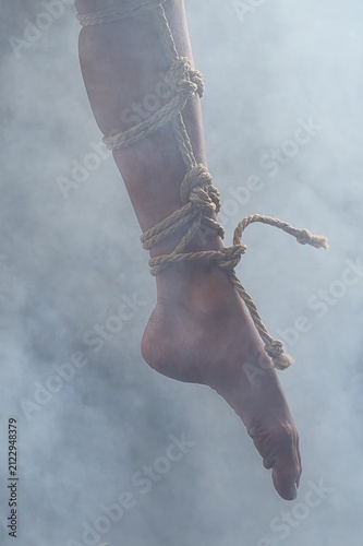 Woman's leg and foot bound with intricate shibari ropes against a smoky, gray studio background.