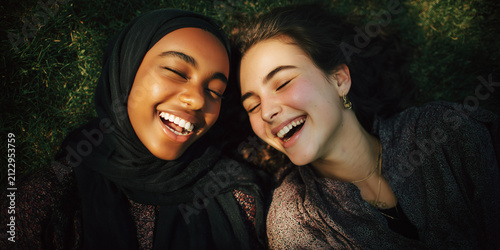 Two young women of diverse backgrounds lying on the grass, laughing heartily. Their eyes are closed in pure joy and happiness, sharing a moment of genuine connection