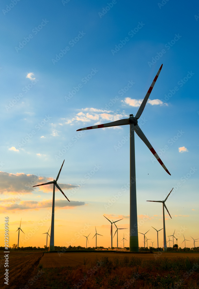 Fototapeta premium Wind turbines against sunset sky. Wind turbines stand tall in a field during sunset with colorful clouds in the sky and fields in the foreground.