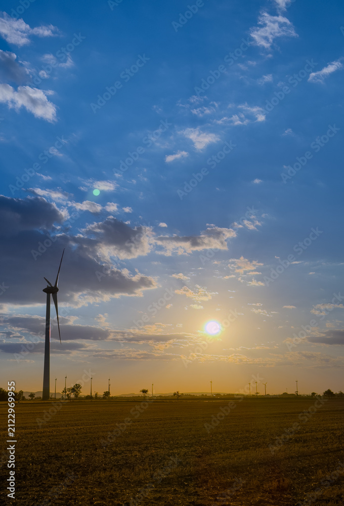 Obraz premium Sunset over a field with wind turbine. A wind turbine stands tall in a field as the sun sets in the background. Clouds fill the sky during the evening.