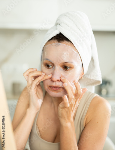 Foto Serene woman with towel head wrap looking in mirror and gently placing cosmetic face mask on, enjoying moment of tranquility and self-care