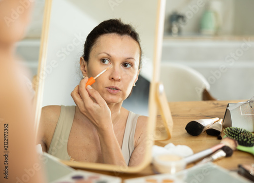 фотографія Serene confident adult woman applying under-eye concealer carefully in well-lit bedroom, sitting before wooden framed makeup mirror