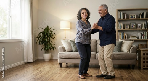 Senior Hispanic couple dancing and laughing in their living room. Older man and woman holding hands and enjoying time together at home. Emotional wellness and active aging concept