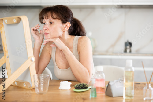 Картината върху платно Adult woman sitting at table and inspecting face in mirror, engaging in regular skincare practice at home