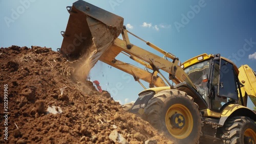 Heavy machinery at work on a construction site with a yellow backhoe loader dumping a bucket of soil onto a large mound of earth under a clear blue sky during earthmoving operations