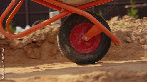 Low angle perspective of a construction worker pushing an orange wheelbarrow filled with sand, the single wheel digging into the soft ground while moving across the building site