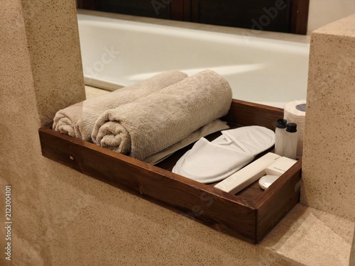 Rolled towels and slippers on a wooden tray beside a hotel bathtub