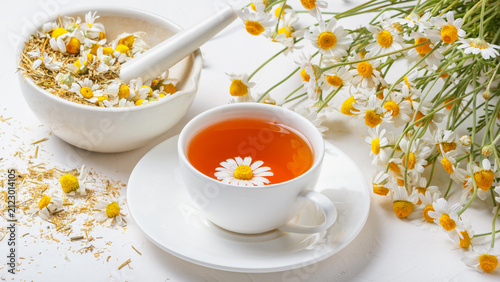 White cup of hot chamomile tea, a mortar with dry daisies buds and a bouquet of Matricaria chamomilla on concrete background. Herbal drink, healthy lifestyle concept.