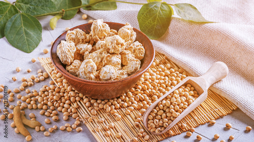 Textured soy protein chunks (nutri nuggets), raw soy meat for vegans in clay bowl on a bamboo napkin against the background of burlap and branch young green pods soybean, closeup with selective focus