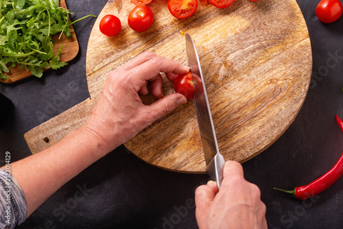 The chef slicing fresh tomatoes for a salad using a large kitchen knife on a wooden chopping board, top view
