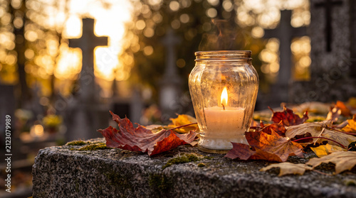 A glowing memorial candle placed on a cemetery headstone surrounded by vibrant autumn leaves, with blurred crosses in the background at sunset.