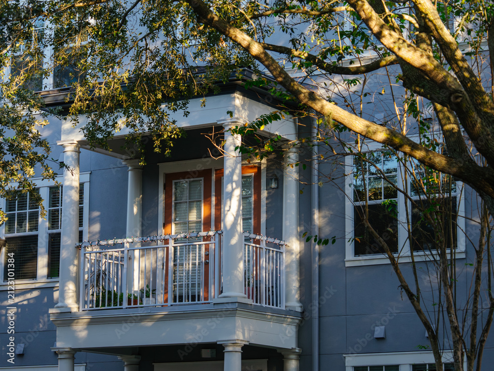 Fototapeta premium A contemporary apartment building features a balcony framed by leafy trees and warm sunlight. The elegant residential architecture creates a peaceful urban living environment.
