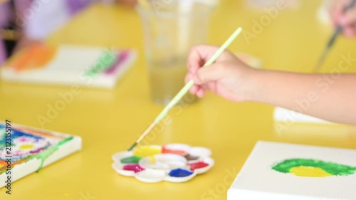 Close up of a child's hand holding a paintbrush over a colorful paint palette on a yellow table during an art class.