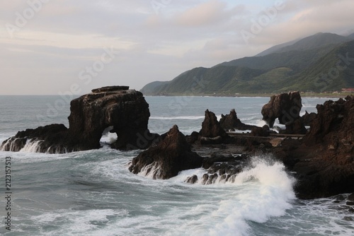 Dramatic rocky coastal sea stacks with crashing white waves against dark volcanic rocks, lush green mountain ridges, and overcast Pacific horizon in Taiwan