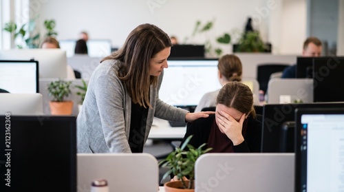 A colleague comforting another colleague in an office environment. One woman has her hand on the other's shoulder as she cries at her desk, surrounded by computers and office supplies