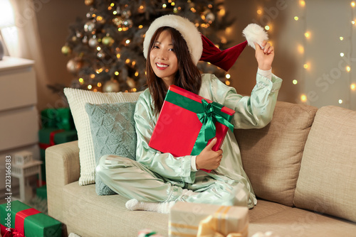 Beautiful young happy Asian woman in Santa hat with Christmas gift box sittin...