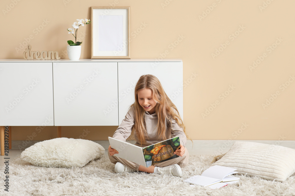 Fototapeta premium Little girl reading book while sitting on floor at home