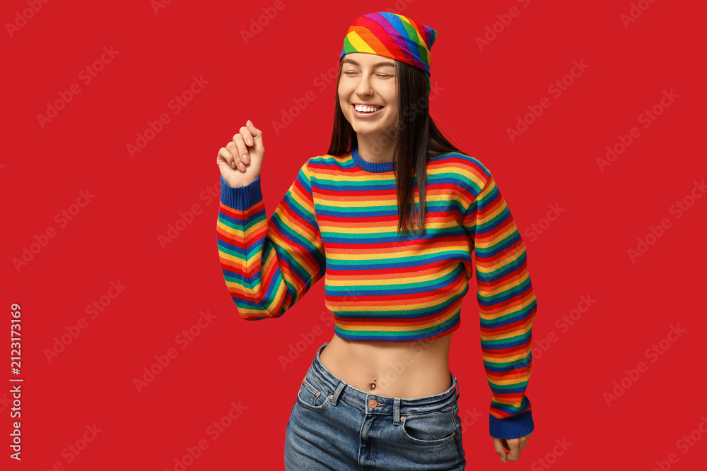 Fototapeta premium Beautiful young happy woman in rainbow sweater with piercing navel on red background