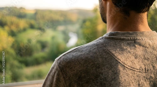 Man gazes out a large window at a lush green valley during the golden hour of a peaceful afternoon.