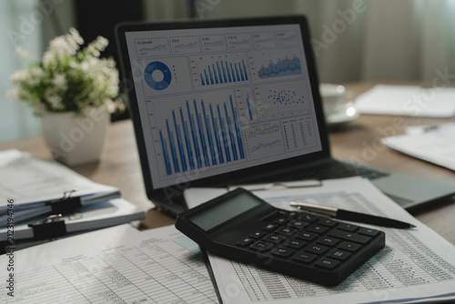 Laptop displays charts and metrics beside a calculator resting on printed spreadsheets and a pen, showing a paperwork-based data analysis workflow for reporting and financial review.