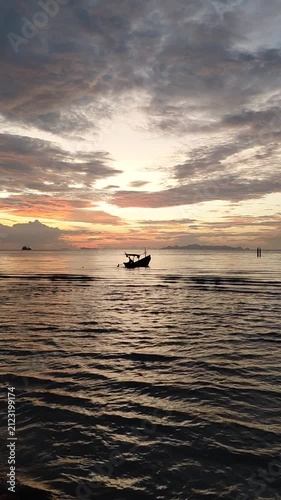 A small wooden fishing boat silhouette floating gently on gentle ocean waves during a vibrant golden hour sunset in Thailand. The sky is filled with dramatic clouds glowing in orange. Thailand.