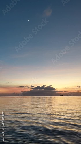 Vertical view of a calm ocean at dusk or dawn, featuring a gentle twilight sky gradient, subtle golden sunlight reflections on water waves, clouds on the horizon, and a visible crescent moon.