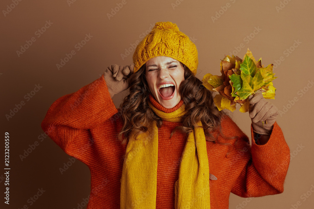 Obraz premium A woman in a yellow knit hat, orange sweater, and scarf playfully shouts with eyes closed, fingers in ears, holding autumn leaves. Against a brown background, she joyfully reacts to the fall season.