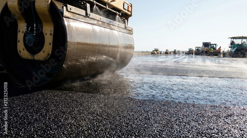 Road roller compacting fresh asphalt layer with water spray on new construction site, infrastructure development.