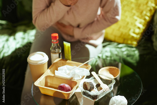 Clutching her stomach at a table filled with fast food, a woman experiences indigestion. The sushi and sauce containers suggest a quick, unhealthy meal that led to physical distress.