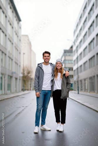 Smiling couple stand side by side on an empty urban street. Buildings line the path, and the weather appears cloudy, creating a calm atmosphere. Generative AI.
