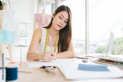 Fashion designer in a bright studio sketches clothing designs at a desk, measuring tape around neck, mannequin and fabrics in the background.