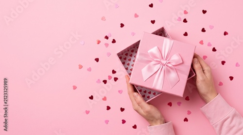 Hands opening a pink gift box with a bow surrounded by heartshaped confetti on a pink background
