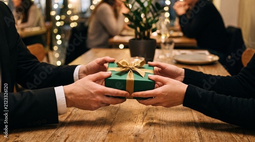 Closeup of a couple exchanging a green gift box with a gold ribbon at a wooden table in a restaurant