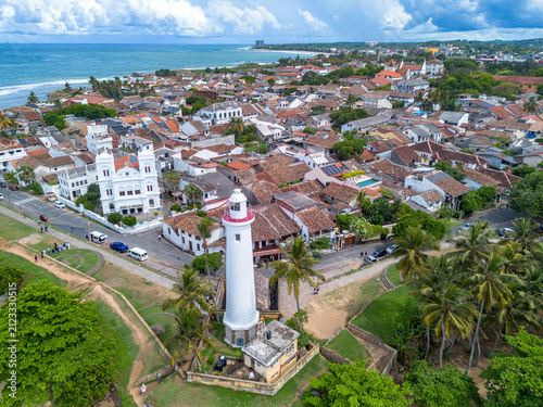 High-angle aerial view of the historic white Galle Lighthouse standing along the stone ramparts next to the Meeran Jumma Mosque and terracotta roofed buildings in Galle Fort, Sri Lanka.