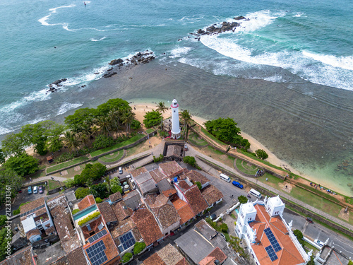 High-angle top-down drone shot of the Galle Lighthouse standing on Point Utrecht Bastion, surrounded by stone fort walls, sandy beach, and clear turquoise shallow waters. 