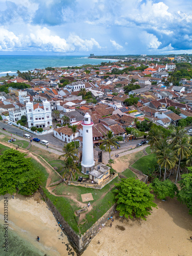 Vertical aerial perspective focusing on Point Utrecht Bastion and Galle Lighthouse, showing tourists walking on the green grass ramparts surrounded by palm trees and colonial architecture.