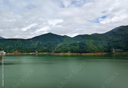 Wide panoramic view of the calm green water at Maojiacun Reservoir surrounded by lush mountain ranges under a cloudy sky in Huize, Yunnan, China.