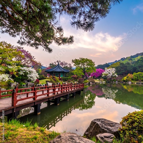 A serene Asian-style garden with a wooden bridge over a pond, surrounded by trees and flowers