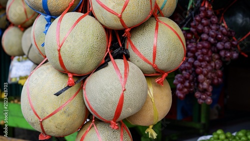 Quadro su tela Traditional Asian market fruit display featuring fresh net-bound cantaloupes and bunches of red grapes ready for retail sale