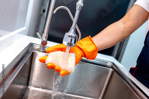 Foto Housekeeper hand wearing orange rubber glove washing sponge under running water from kitchen sink faucet