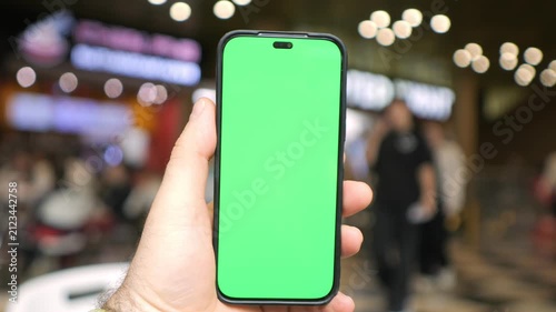 Hand holds smartphone with green screen. Blurred background features warm lights and people. Phone remains steady in second frame. Background shows glowing mall signs and moving figures