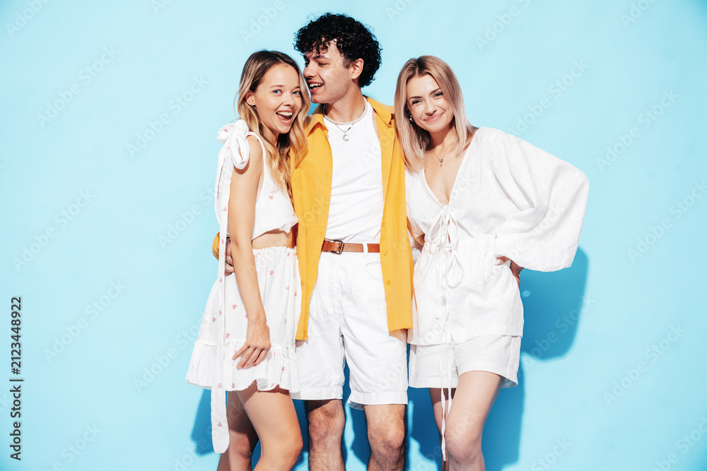 Fototapeta premium Group of young three stylish friends posing in studio. Fashion man and two cute female dressed in casual summer clothes. Smiling models having fun. Cheerful women and guy near blue wall