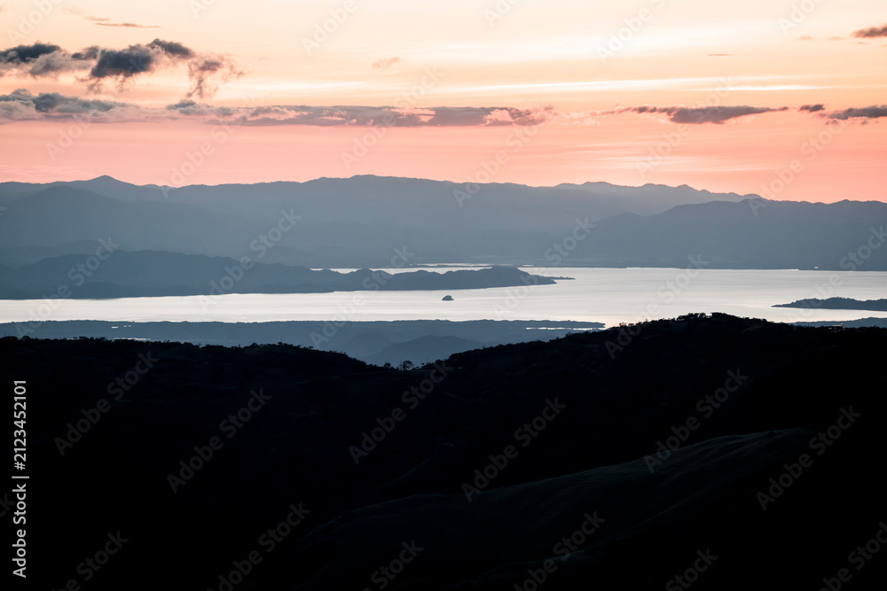 Fototapeta premium Panoramic layered mountain and lake landscape at sunset with silhouetted hills and pastel sky
