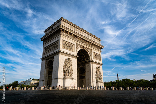 The Arc de Triomphe under a blue summer sky in Paris, France, with the Eiffel Tower visible in the distance and tourists gathered at its base
