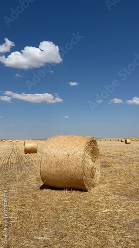 A cinematic 4K low-angle tracking shot of a large round hay bale in a golden harvested wheat field under a bright blue summer sky with scattered white clouds.