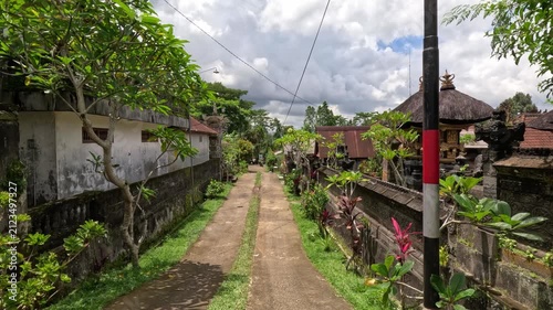 Wide shot of a residential street on Bali island during a sunny day, showing houses with dedicated Balinese shrines, lush greenery, and a clear blue sky.