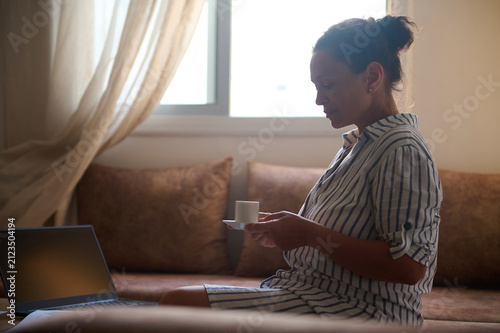 Woman drinking coffee while working on laptop at home in a relaxed morning routine