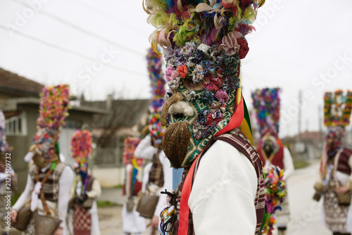 Grebentsi kukeri performer with rooster-skin beard, horse-tail hair, decorated headpiece, ribbons and bells.