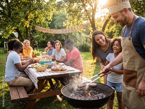 Man grilling meat at outdoor summer backyard barbecue party with diverse friends and family gathering in garden